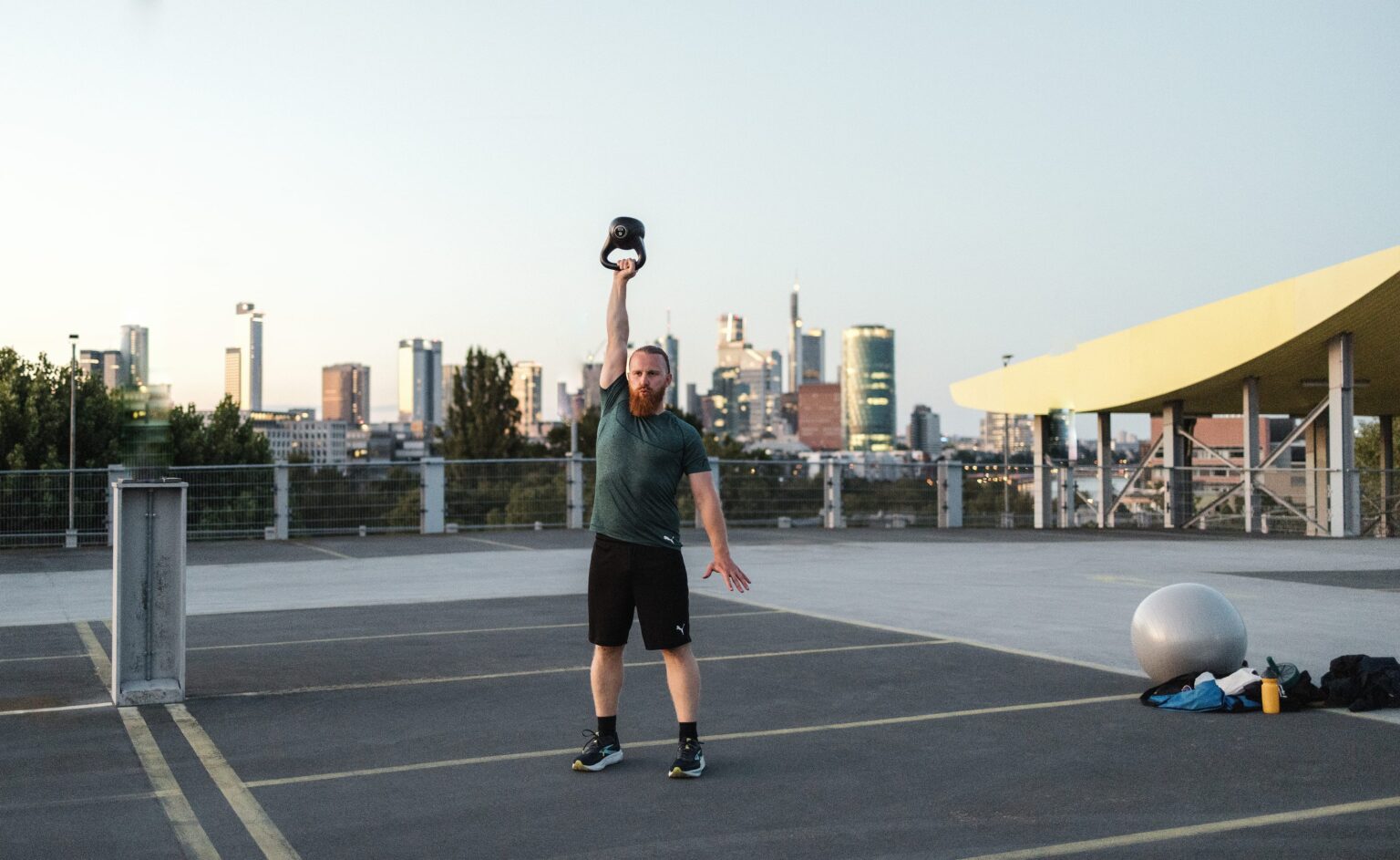 Personal Trainer Matthäus drückt eine Kettlebell über Kopf auf einem Parkdeck, dahinter die Hochhäuser von Frankfurt