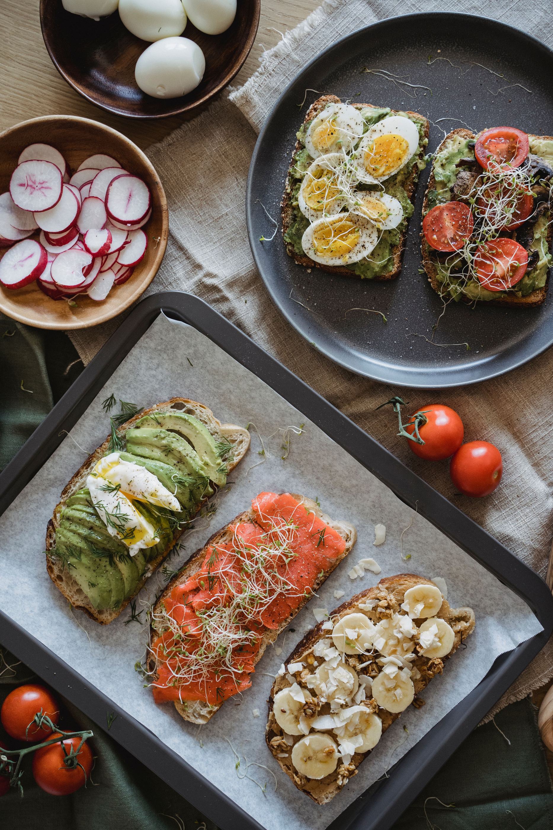Vier belegte Vollkornbrote mit Avocado, Ei, Lachs und Banane, dazu Radieschen und Tomaten — Beispiel für alltagstaugliche Ernährung