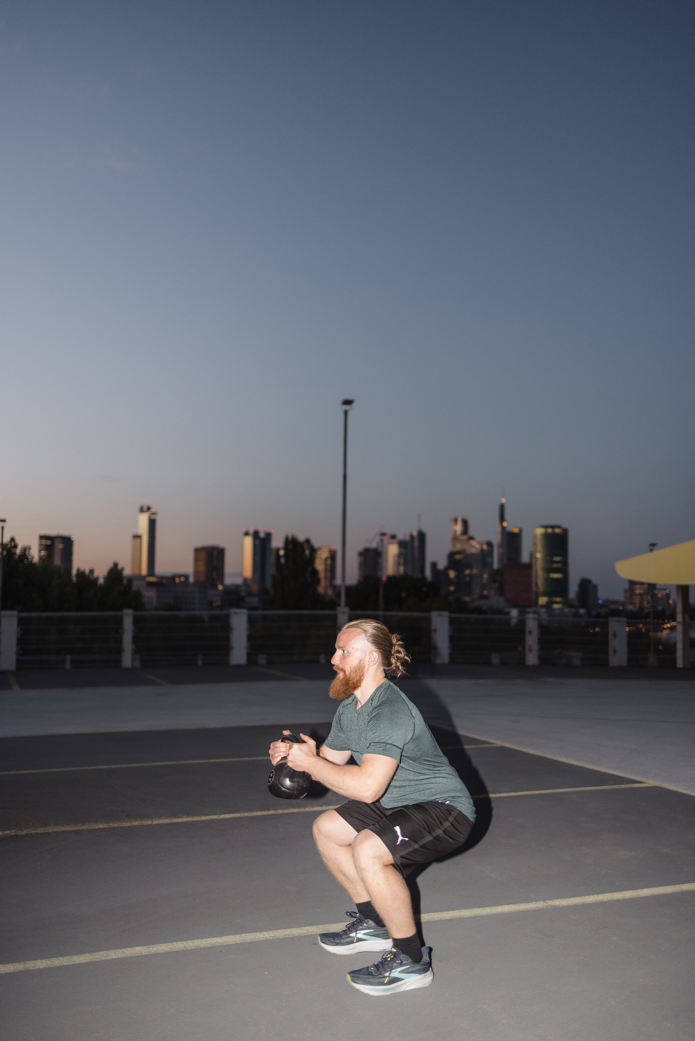 Personal Trainer Matthäus in der tiefen Kniebeuge mit Kettlebell vor der Brust, im Hintergrund die beleuchtete Skyline von Frankfurt in der Abenddämmerung