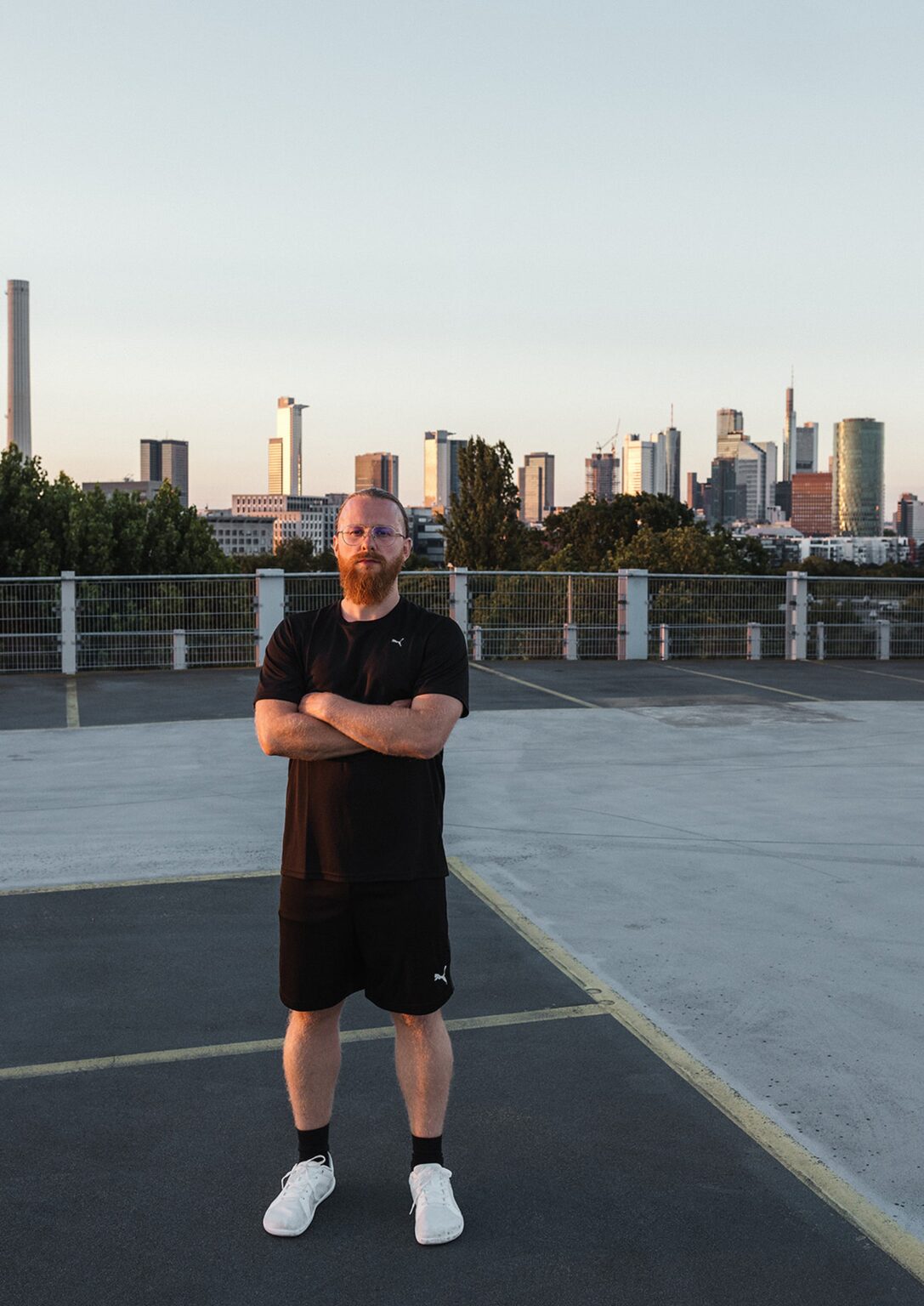 Personal Trainer Matthäus steht mit verschränkten Armen auf einem Parkdeck vor der Frankfurter Skyline