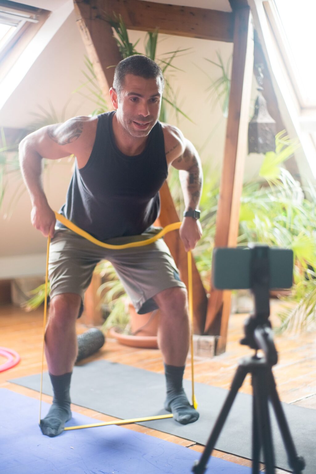 Mann trainiert zu Hause mit einem Widerstandsband und filmt sich dabei mit dem Handy auf einem Stativ — Training per Videocall